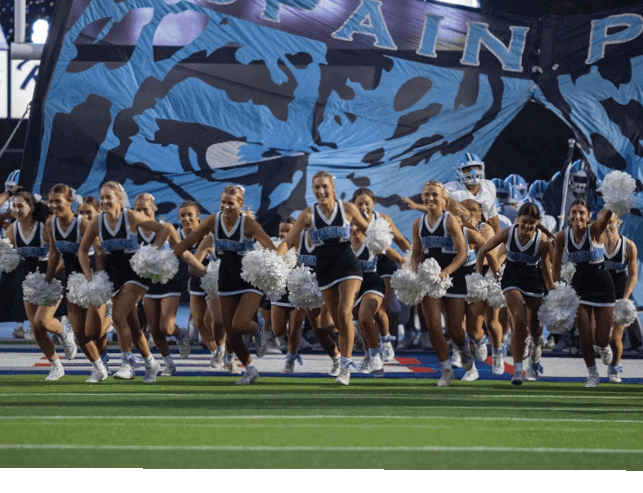 A group of cheerleaders in matching uniforms with pom-poms are running forward on a football field in front of a large banner. Description generated by AI