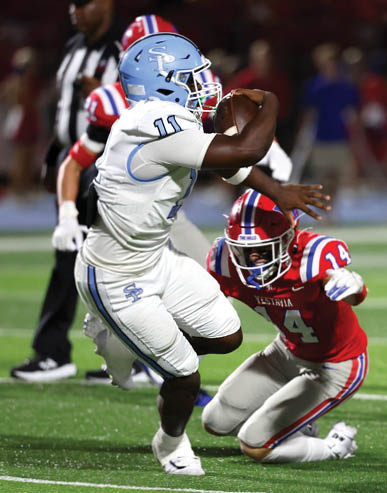 An adult in a white and blue football uniform is holding a football while another adult in a red and white football uniform is crouching and reaching toward them on a green field. Description generated by AI