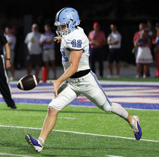 A person wearing a light blue helmet and white football uniform with the number 42 is kicking a football on a green field with spectators in the background. Description generated by AI