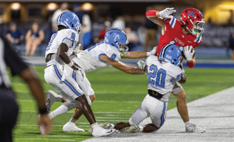 A person in a red football uniform is running with the ball while three people in white and blue uniforms attempt to tackle them on a football field. Description generated by AI