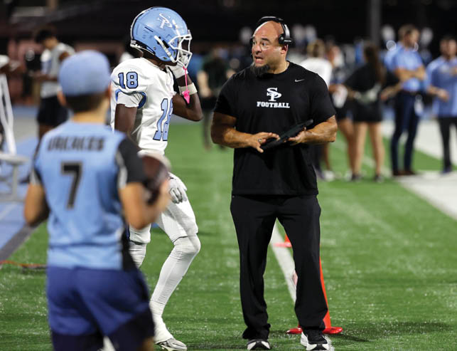 A person wearing a headset and black sportswear is holding a tablet and talking to a person in white football gear with the number 18 on the jersey while another person in light blue sportswear with the number 7 is in the foreground on a football field. Description generated by AI