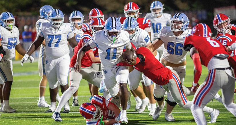 A group of people in football uniforms are actively engaged in a game on a field with one person in white running with the ball while others in red attempt to tackle or block. Description generated by AI