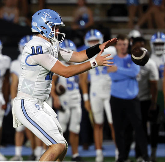 A person wearing a blue helmet and white football uniform with the number 10 is reaching out to catch a football during a game with other players and a coach in the background. Description generated by AI