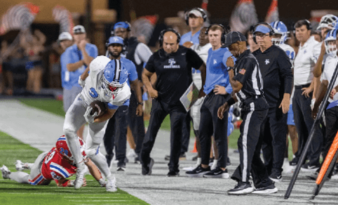 A person in a white and blue football uniform is running with the ball while another person in a red and blue uniform is on the ground attempting to tackle them near the sideline with coaches and officials watching nearby. Description generated by AI