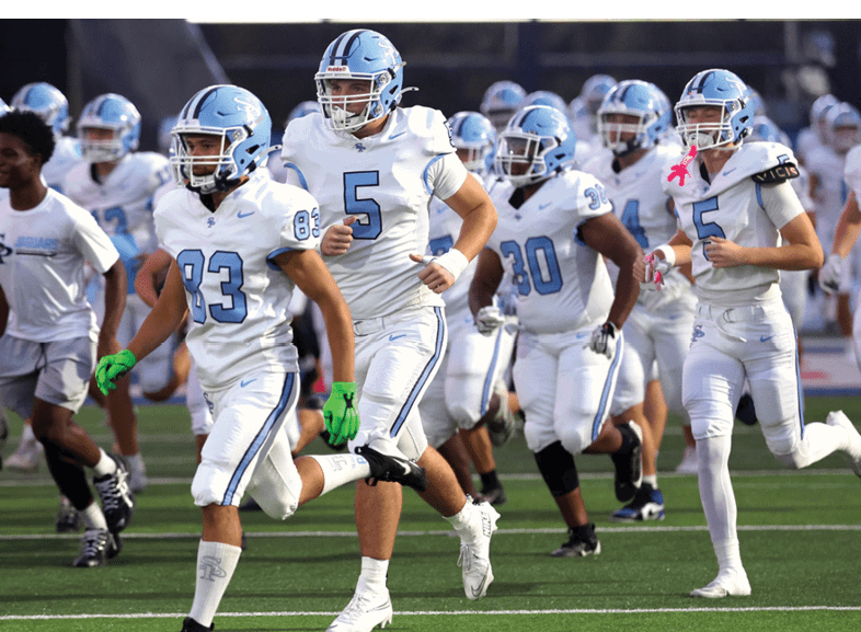 A group of people wearing white football uniforms and helmets are running on a green field during a game or practice. Description generated by AI
