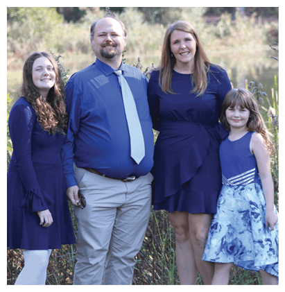 Four people dressed in blue clothing stand closely together outdoors with greenery in the background, smiling and posing for the photo. Description generated by AI