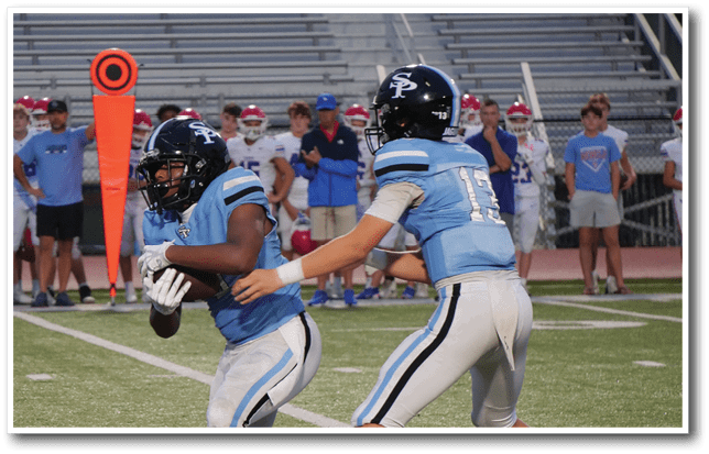Two football players in blue and white uniforms are on the field with one holding the ball and the other reaching out to support, while people stand in the background near the sideline. Description generated by AI