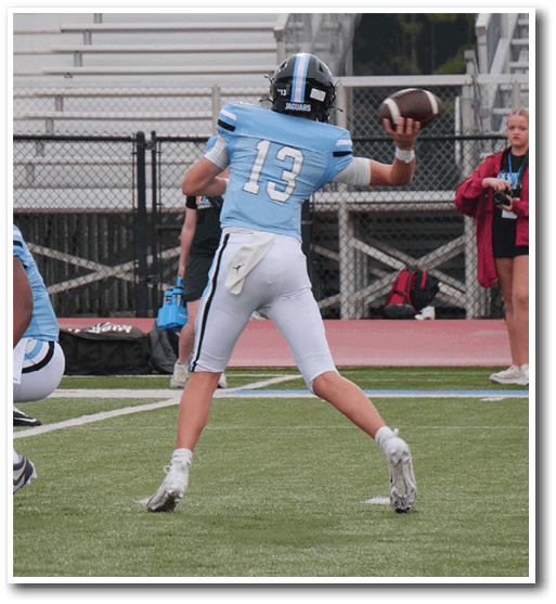 A person wearing a light blue football jersey with the number 13 is preparing to throw a football on a field with two people standing near an orange down marker in the background. Description generated by AI