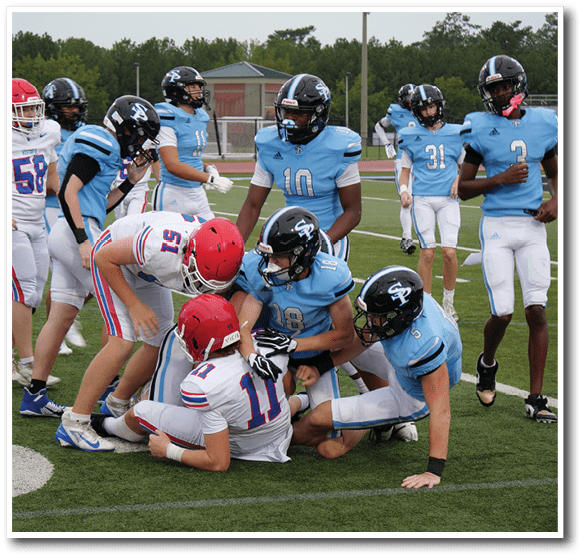 A group of people wearing football uniforms are gathered on a field with some standing and others crouching or leaning in close. Description generated by AI