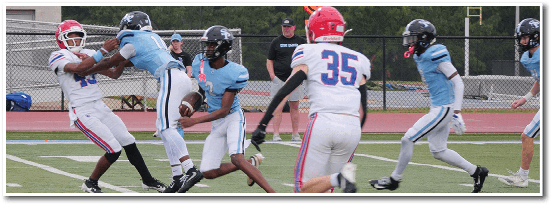 People in football uniforms are actively playing on a field with one person tackling another while others run nearby and a person walks along the sideline. Description generated by AI