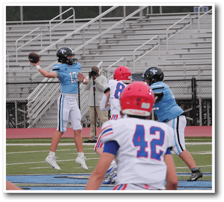 A group of people in football uniforms are on a field with one person in a blue jersey preparing to throw a football while others in red and blue jerseys are positioned around. Description generated by AI