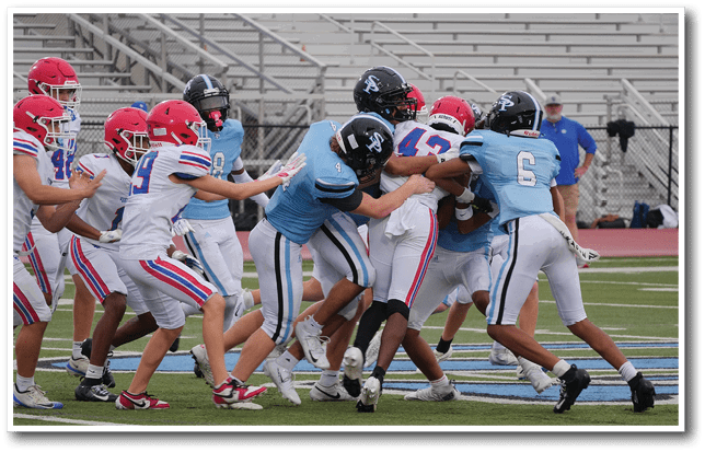 A group of people wearing football uniforms are engaged in a tackle on a football field with empty bleachers in the background. Description generated by AI