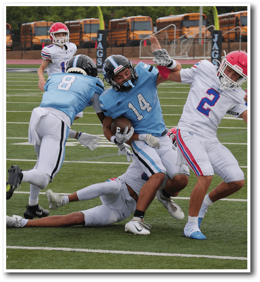 A group of people in football uniforms are actively engaged in a play on a football field with one person holding the ball while others attempt to tackle or block. Description generated by AI