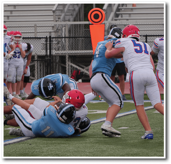 People wearing football uniforms are engaged in a play on a football field with some players blocking and others preparing to move. Description generated by AI