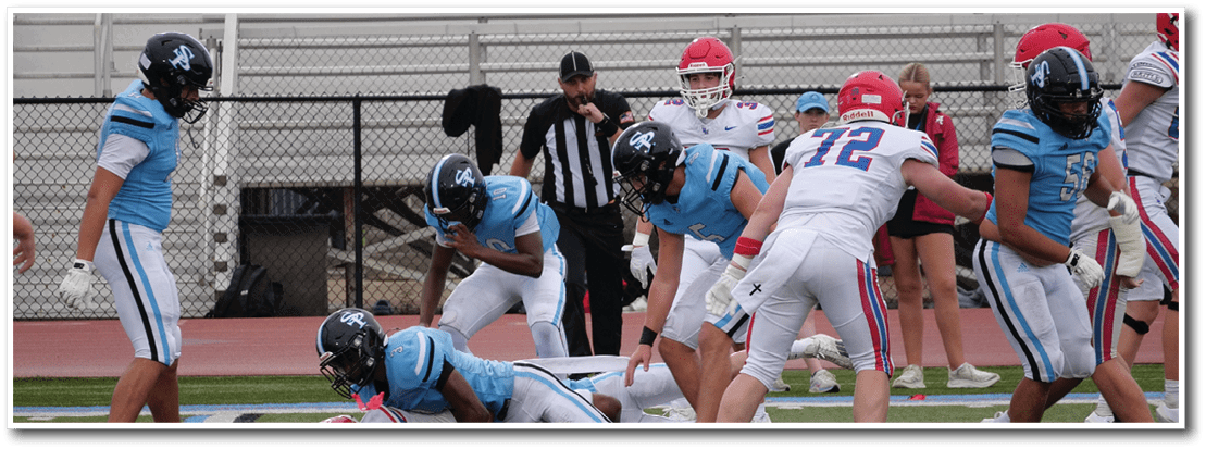 Several people in football uniforms are on a field with one person lying on the ground while others stand or crouch nearby, and a referee and cheerleaders are visible in the background. Description generated by AI