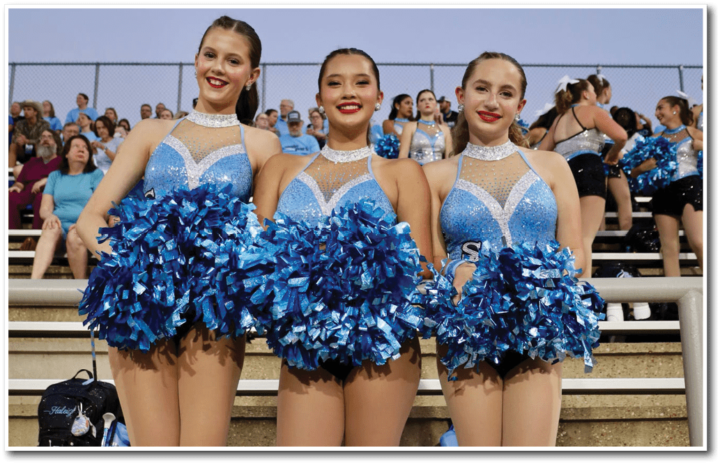 Three people wearing blue and white dance costumes smile in front of a crowd at an outdoor event under a clear sky. Description generated by AI