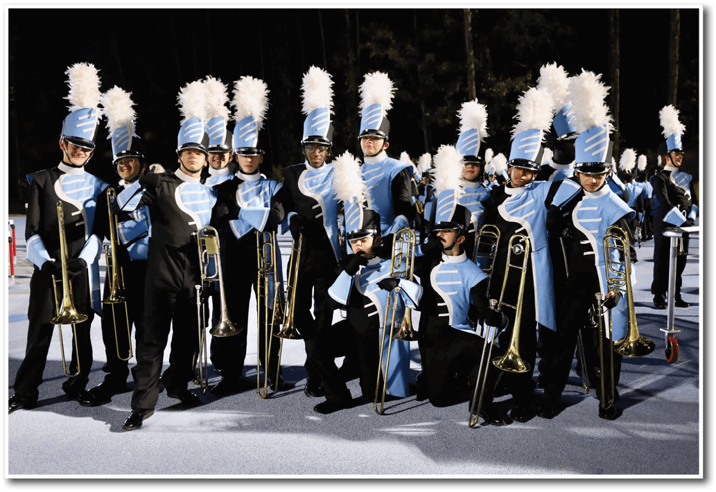 A group of people dressed in blue and black marching band uniforms with tall white plumes on their hats are standing and kneeling on a field holding trombones. Description generated by AI