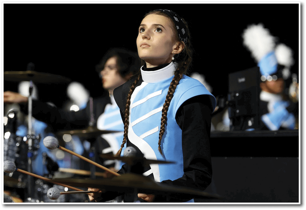 A person in a blue and black marching band uniform with braided hair is playing percussion instruments with mallets during a nighttime performance. Description generated by AI