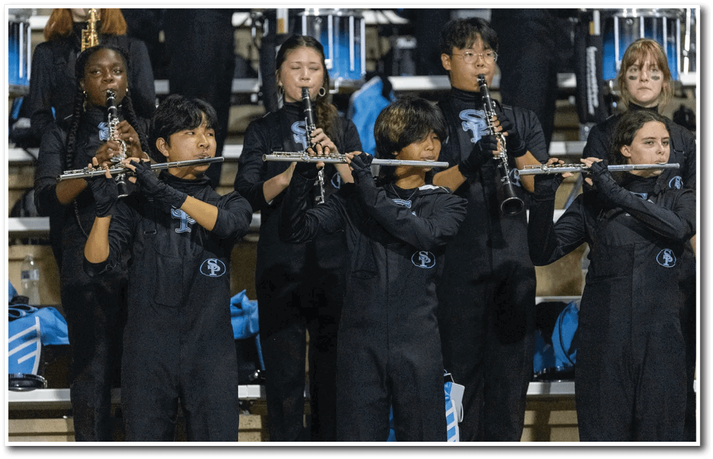 People wearing matching dark uniforms are playing flutes and clarinets while standing in rows on bleachers. Description generated by AI
