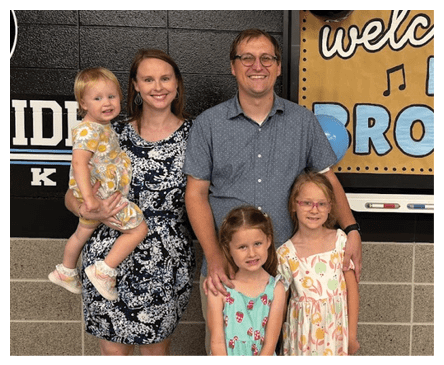 A group of five people, including three children in colorful dresses and two adults, stand together smiling in front of a wall with a sign and markers. Description generated by AI