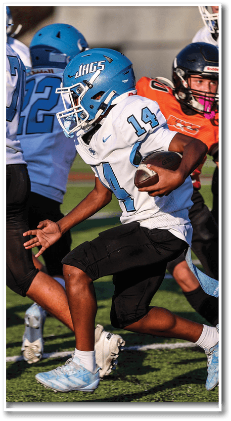 A person wearing a blue helmet and white football jersey with the number 14 is running with a football while other players in blue and orange uniforms are nearby on a grassy field. Description generated by AI