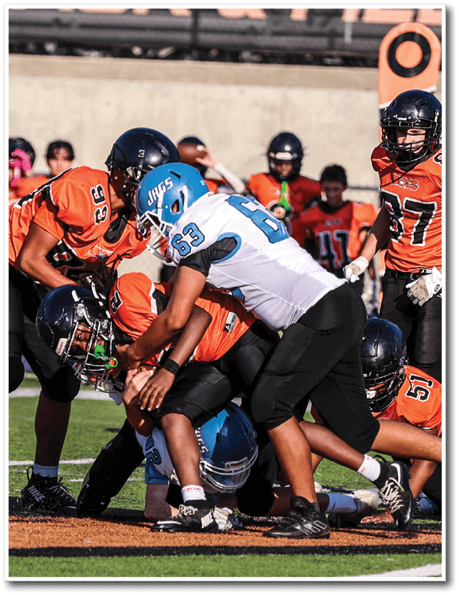 Several people wearing football helmets and uniforms are engaged in a play on a football field with some players in orange and one in white and blue. Description generated by AI