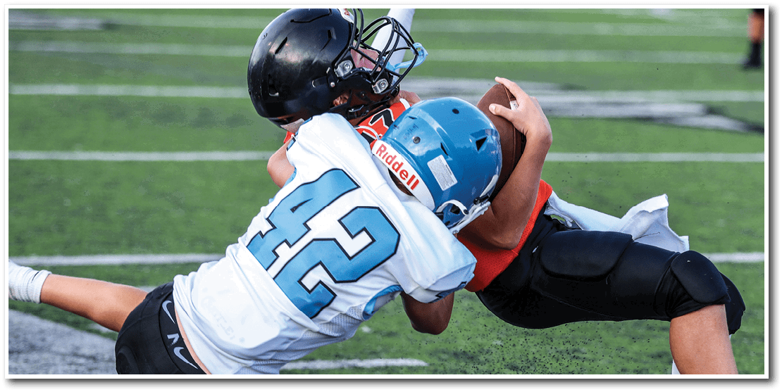Two people in football gear are engaged in a tackle on a grassy field near a marked boundary line. Description generated by AI