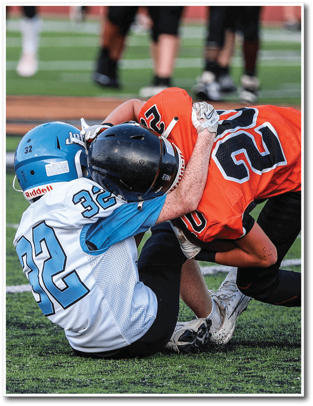 Two people wearing football helmets and uniforms are engaged in a tackle on a grassy field during a game. Description generated by AI