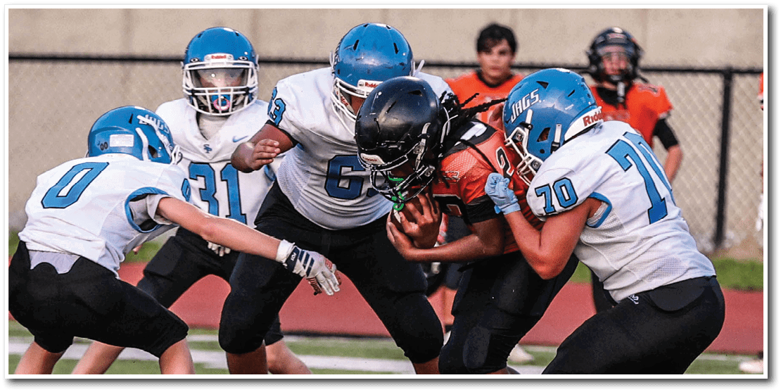 Four people in football uniforms are engaged in a play on a field with one person in black holding the ball while three others in white and blue helmets attempt to tackle. Description generated by AI