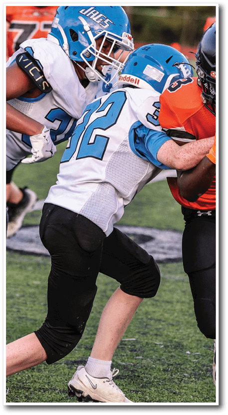 A person in an orange and black football uniform is running with the ball while being tackled by another person in a white and blue uniform on a grassy field. Description generated by AI