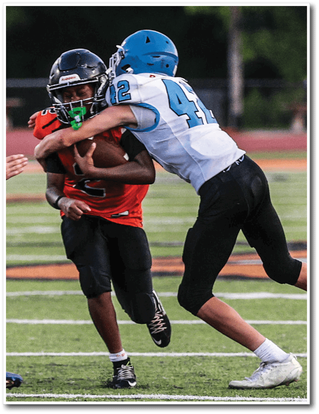 Two people wearing football uniforms are engaged in a tackle on a grassy field during a game. Description generated by AI