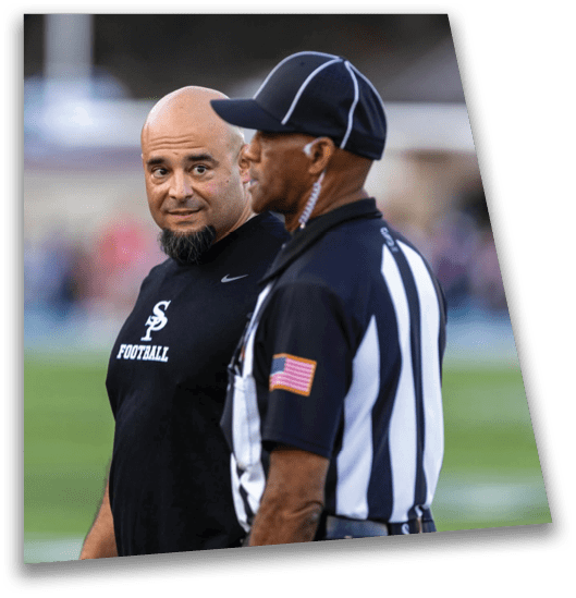 Two adults stand on a sports field, one wearing a black shirt with a football logo and the other dressed as a referee with a striped shirt and an American flag patch. Description generated by AI
