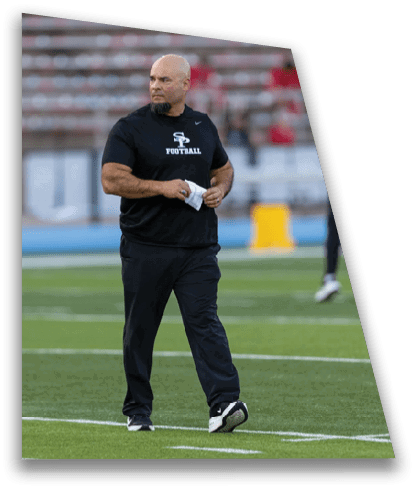 A person wearing a black shirt and pants stands on a football field holding a piece of paper while other people in sports attire are visible in the background. Description generated by AI