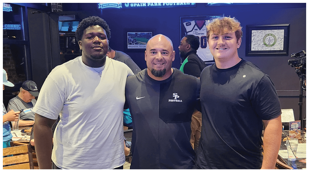 Three adults stand closely together indoors with one wearing a white shirt, another in a black sports shirt with a football logo, and the third in a plain black shirt, all smiling. Description generated by AI
