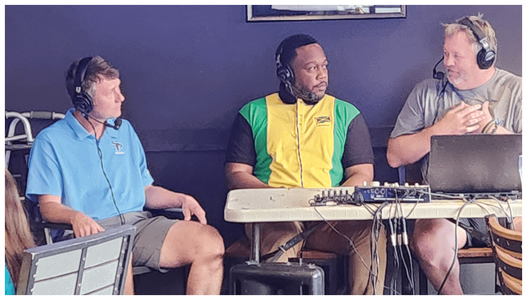 Three adults wearing headsets are seated and engaged in a discussion around a table with audio equipment and a laptop. Description generated by AI