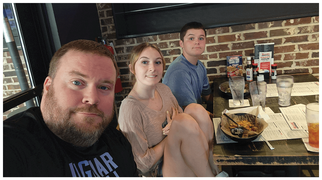 Three adults sitting closely together at a restaurant table with food and drinks, smiling at the camera. Description generated by AI