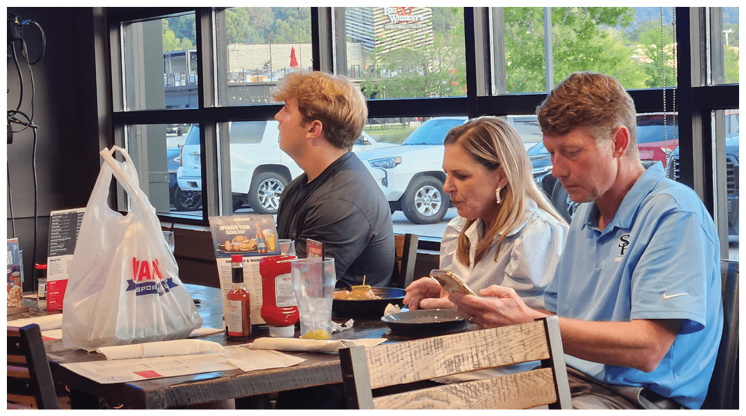 Three adults sit side by side at a restaurant counter with food and drinks in front of them, one looking at a phone while the others face forward. Description generated by AI