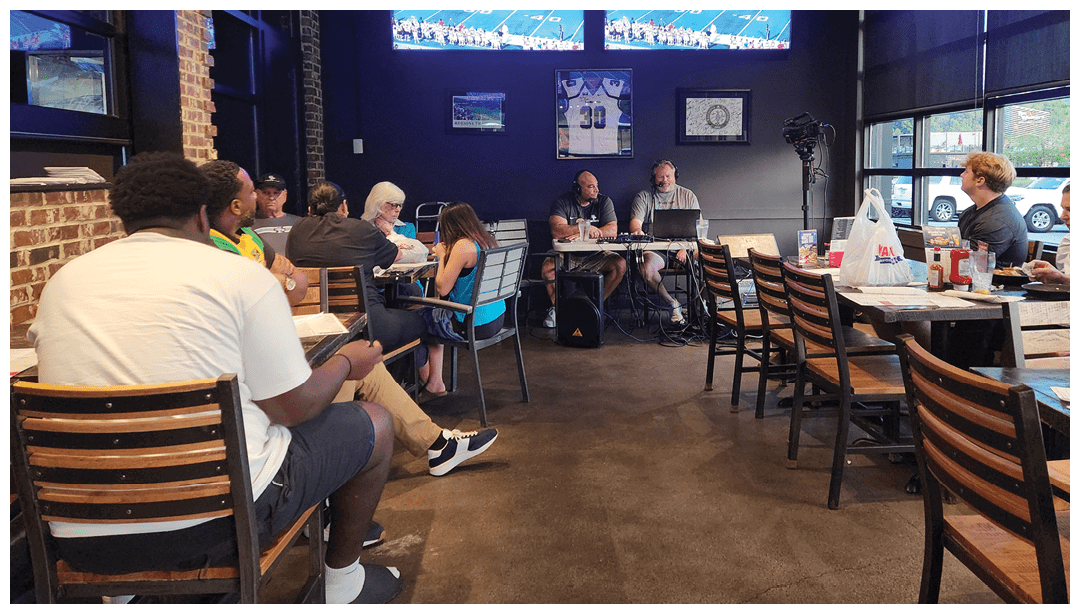 Adults sit in a casual dining area watching a sports game on two large wall-mounted TVs while two people speak into microphones at a table in front of them. Description generated by AI