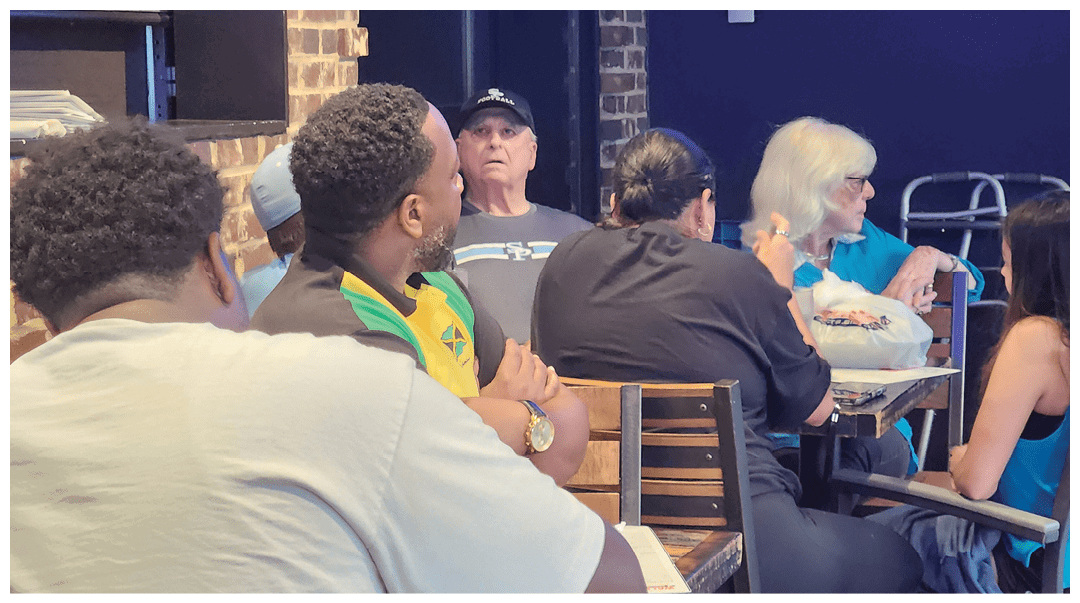 Several people are seated indoors around tables, engaged in conversation, with one person wearing a cap and another with light-colored hair near a walker. Description generated by AI