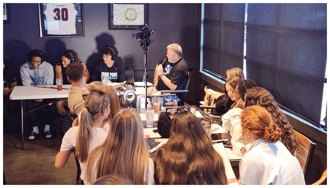 A group of people with headphones are seated at a table speaking into microphones while an audience listens attentively in a room with dark curtains. Description generated by AI