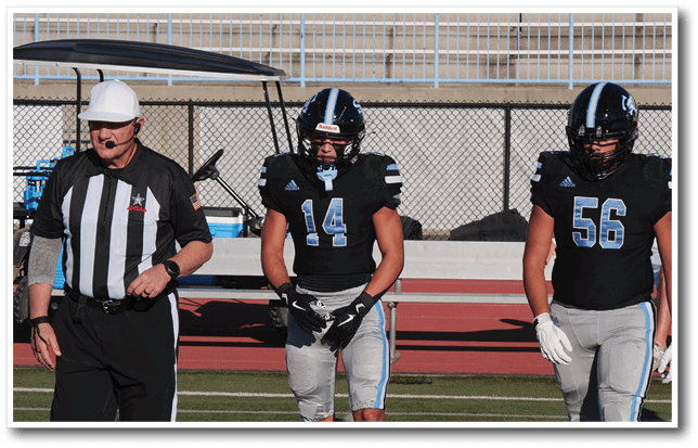 A referee wearing a white hat and striped shirt stands next to two football players in black jerseys and helmets on a sports field. Description generated by AI