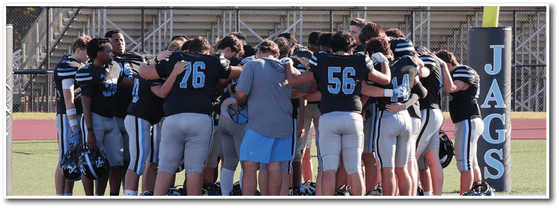 A group of people wearing football uniforms and one person in casual clothing are huddled together on a sports field near a goal post with “JAGS" written on the padding. Description generated by AI
