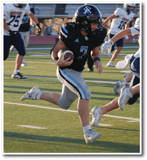 A person wearing a black football uniform with the number 7 is running on a field while holding a football, pursued by others in white and blue uniforms. Description generated by AI