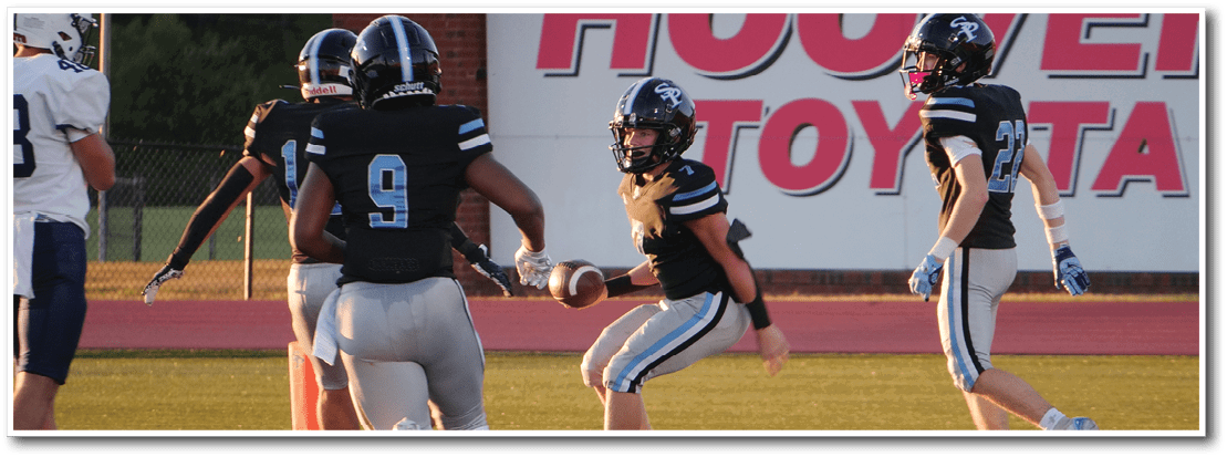People wearing football uniforms are on a field near the end zone, with one person holding a football and others standing nearby. Description generated by AI