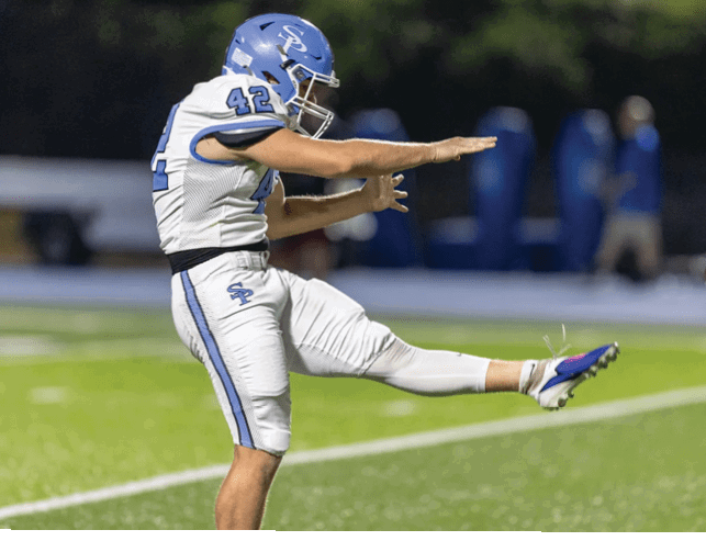 A person in a blue and white football uniform is kicking a ball on a grassy field during a game. Description generated by AI