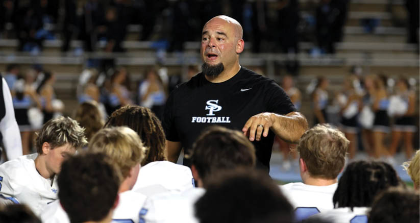 An adult wearing a black shirt with a football logo is speaking to a group of people in white sports uniforms on a field with blurred spectators in the background. Description generated by AI