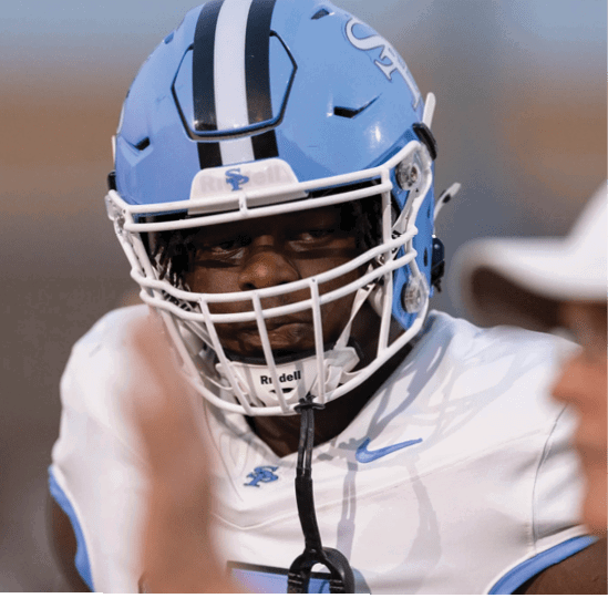A person wearing a light blue football helmet and white jersey is focused and appears to be preparing for a play. Description generated by AI