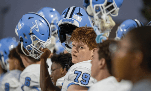 A group of people wearing blue and white football helmets and uniforms are gathered closely, with one person in the center looking directly at the camera. Description generated by AI
