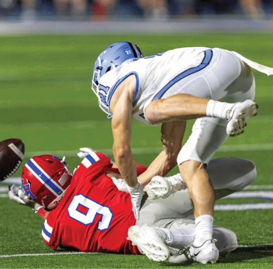 Two people in football uniforms are engaged in a tackle on a grassy field during a game. Description generated by AI