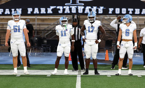 Four adults in white football uniforms stand on a field with a referee behind them and a banner reading “PAIN PARK JAGUARS FOOTBALL" in the background. Description generated by AI